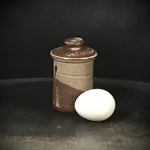 May include: A brown and white ceramic jar with a lid, sitting on a dark surface next to a white egg.