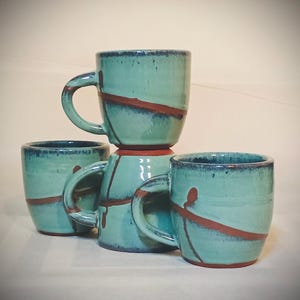 May include: Four handmade ceramic mugs in teal and brown. Each mug has a unique design with a teal glaze and a diagonal brown stripe. The mugs are stacked and arranged to showcase their individual shapes and details.