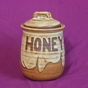 May include: A ceramic honey jar with a lid. The jar is beige with brown accents and the word "HONEY" in large, brown letters. The bottom of the jar has a brown, dripped honey design. The lid has a small handle.