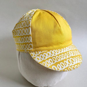 May include: A yellow cycling cap with a white patterned design. The cap has a curved brim and a rounded crown.