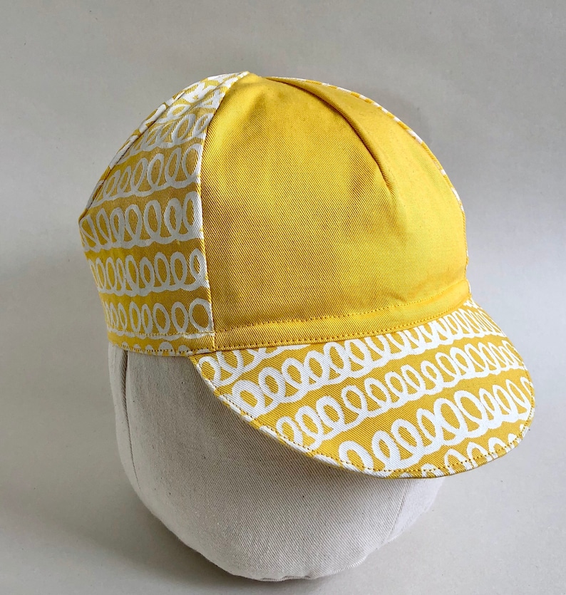 May include: A yellow cycling cap with a white patterned design. The cap has a curved brim and a rounded crown.