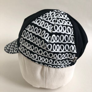May include: Black cycling cap with a white pattern of repeating loops. The cap has a short bill.