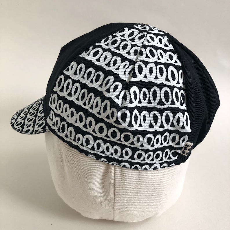 May include: Black cycling cap with a white pattern of repeating loops. The cap has a short bill.