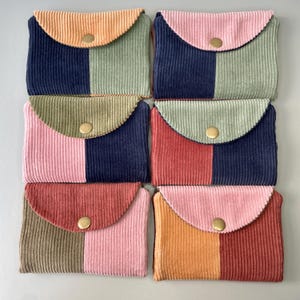 May include: A selection of corduroy wallets in shades of pink, orange, green, and navy blue. Each wallet has a two-tone design and a flap closure with a gold-coloured snap. The wallets are arranged in a grid pattern.