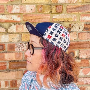 May include: A navy blue baseball cap featuring a white and red pattern with Japanese characters. The cap has a curved peak and a white interior. The cap is worn by a person with glasses and pink and orange hair.