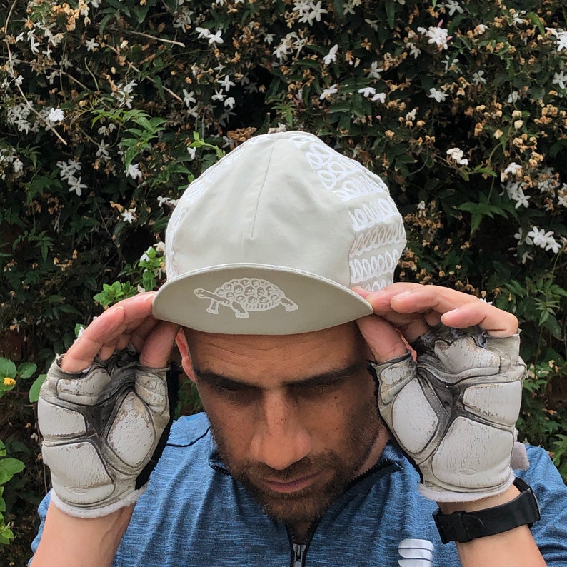 May include: A gray cycling cap with a white embroidered turtle design. The cap has a white patterned brim.