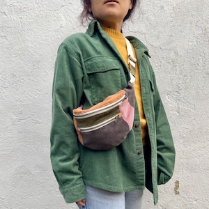 May include: A green corduroy shirt with a multi-coloured corduroy fanny pack worn over the shoulder. The fanny pack has a zipper closure and is made of a patchwork of different coloured corduroy fabrics.