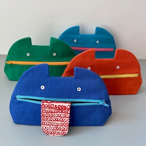 May include: Four colourful zippered pouches with a whimsical cat face design. The pouches are green, blue, orange, and teal. The blue pouch has a red and white patterned fabric lining.