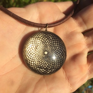 Könnte beinhalten: Eine goldene und schwarze geometrische Anhänger-Halskette mit einem Lederband. Der Anhänger hat ein kreisförmiges Design mit einem sich wiederholenden Muster aus Dreiecken.