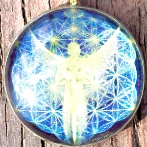 May include: Round pendant with a gold-colored bail. The pendant features a blue and white design with an angel figure and geometric patterns. The pendant is set against a wooden background.