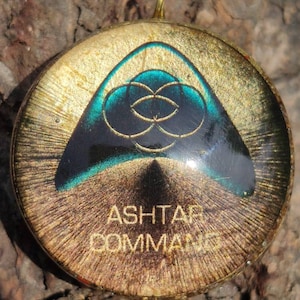 Könnte beinhalten: Ein runder Anhänger in Gold mit einer schwarzen dreieckigen Form, die ein grünes und blaues geometrisches Design enthält. Der Text "Ashtar Command" ist auf den Anhänger gedruckt.