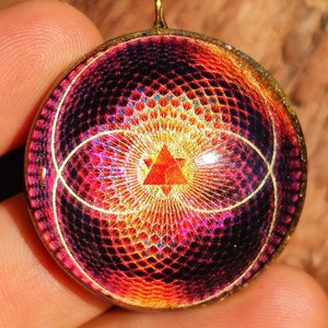 Pode incluir: Um pingente circular com um padrão geométrico vibrante e multicolorido em tons de roxo profundo, magenta e laranja brilhante. No centro, uma pequena estrela vermelha de seis pontas é cercada por linhas radiantes.