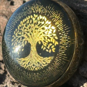 Puede incluir: Colgante redondo con un diseño dorado de árbol de la vida sobre fondo negro. El colgante tiene un marco de metal dorado y un lazo para una cadena. El diseño del árbol está detallado con hojas y raíces.