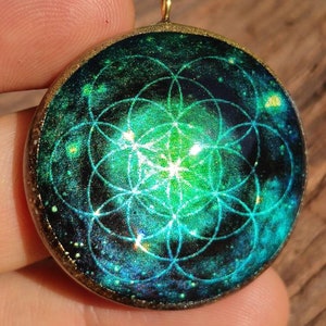 Pode incluir: Um pingente redondo, verde e dourado com um design geométrico dentro. O design é um padrão de flor da vida, um símbolo de geometria sagrada.