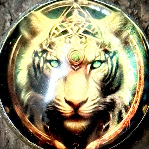 Pode incluir: Um pingente redondo com um tigre branco com olhos verdes. O tigre está usando uma coroa dourada com um design de nó celta. O pingente tem uma borda marrom e dourada.