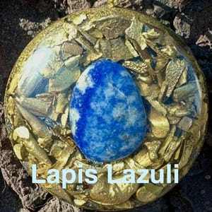 Puede incluir: Un colgante redondo con una piedra de lapislázuli azul en el centro, rodeada de escamas doradas y resina. El texto "Lapis Lazuli" se muestra en la parte inferior. El colgante tiene un lazo en la parte superior para colgar.