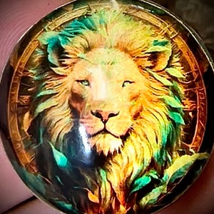 Pode incluir: Um pingente redondo dourado com uma imagem colorida de uma cabeça de leão. O leão tem uma juba verde e amarela e um fundo dourado.