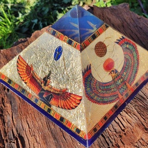 Peut inclure: Une pyramide dorée et bleue avec une image d'une déesse ailée et d'un faucon ailé. La pyramide a un fond doré avec une bordure bleue et un symbole Ankh doré.