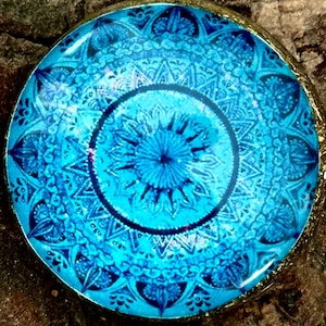 May include: A round pendant with a blue mandala design. The pendant is made of glass and has a gold-colored metal frame.