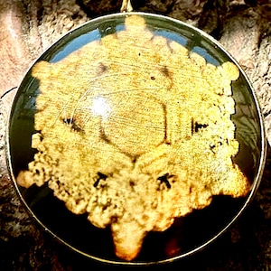 Pode incluir: Um pingente de ouro com um cabochão redondo de resina transparente que apresenta um design detalhado e intrincado em tons dourados e marrons. O design parece ser um padrão floral ou geométrico estilizado.