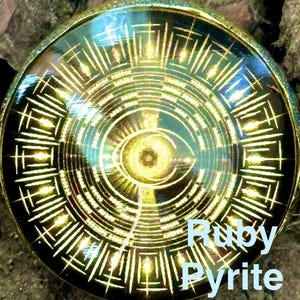Pode incluir: Um pingente circular com uma moldura dourada e um design preto e dourado. O design apresenta um olho no centro, rodeado por padrões geométricos. As palavras "Ruby Pyrite Jade" estão escritas em texto azul claro.