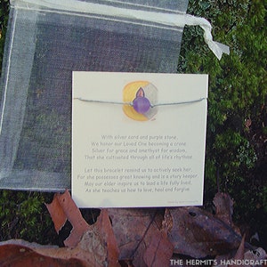 May include: A silver cord bracelet with a purple stone charm. The card reads "With silver cord and purple stone, We honor our Loved One becoming a crone. Silver for grace and amethyst for wisdom, That she cultivated through all of life's rhythms. Let this bracelet remind us to actively seek her, For she possesses great knowing and is a story keeper May our elder inspire us to lead a life fully lived, As she teaches us how to love, heal and forgive. THE HERMIT'S HANDICRAFTS"
