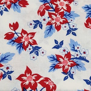 May include: A white fabric featuring a repeating floral pattern. The design consists of large red flowers with pink centers, smaller white blossoms with red centers, and clusters of blue and light blue leaves arranged across the surface.