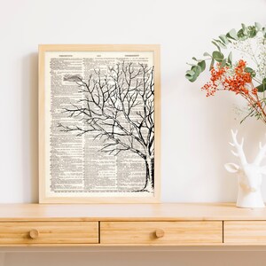 May include: A framed art print featuring a black and white tree illustration. The tree is set against a backdrop of vintage book pages. The frame is a light wood color, and the artwork is displayed on a wooden surface.