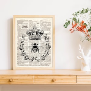 May include: Black and white print of a bee with a crown on top of it, surrounded by a wreath of leaves. The print is on a vintage dictionary page.