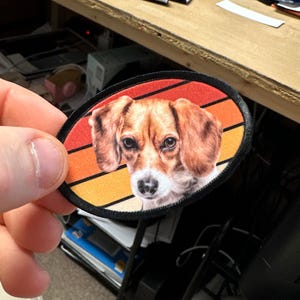 May include: A round, black-edged patch with a retro-style sunset design. The patch features a close-up image of a beagle dog with brown and white fur.
