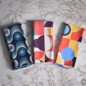 May include: Three colorful towels with geometric patterns. The first towel is blue and white with a swirling pattern. The second towel is red, white, and yellow with a geometric pattern. The third towel is yellow, red, and blue with a geometric pattern.