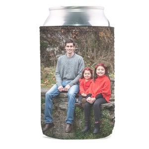 Pu&ograve; includere: Un refrigeratore per lattine con una foto di una famiglia di tre persone. Il padre indossa un maglione grigio e jeans blu. Le due figlie indossano maglioni rossi e hanno i capelli legati con fasce rosse.
