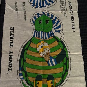 Puede incluir: Un panel de tela con un gráfico impreso de una tortuga llamada Tommy Turtle. La tortuga lleva un gorro y una bufanda de rayas azules y blancas, botas amarillas y sostiene un helado. El texto dice TOMMY TURTLE y CUT ALONG THIS LINE.