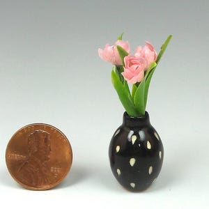 May include: Miniature black vase with white polka dots holding pink flowers and green leaves. A penny is next to the vase for scale. The vase is a handcrafted item.