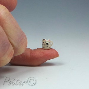 May include: A miniature ceramic mug with a cat face design, held on a fingertip. The tiny mug is white with black spots and a black cat face. The mug has a small handle.