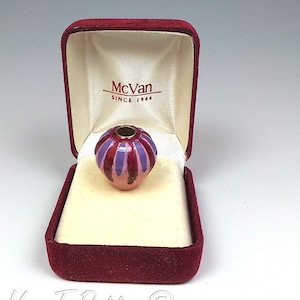 May include: A small, round, hollow bead with vertical stripes in shades of purple and dark red, resting inside a maroon velvet-lined jewelry box. The interior lid of the box features the printed text McVan SINCE 1944 in a dark red font.