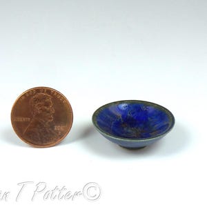 Könnte beinhalten: Eine kleine, runde, blaue Keramikschale mit dunklem Rand. Eine Münze liegt neben der Schale zum Größenvergleich. Das Innere der Schale hat eine strukturierte, glänzende Oberfläche. Der Hintergrund ist weiß.