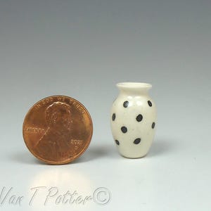 May include: A small, off-white ceramic vase with black polka dots. The vase is next to a penny for size comparison. The vase has a rounded body and a narrow neck. The vase is likely handmade.