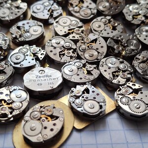 May include: A collection of vintage watch movements, showcasing intricate gears and mechanisms. The silver-toned components are arranged on a light-colored surface, with a few gold-toned bases. One movement has the text "Zentra Container 365".