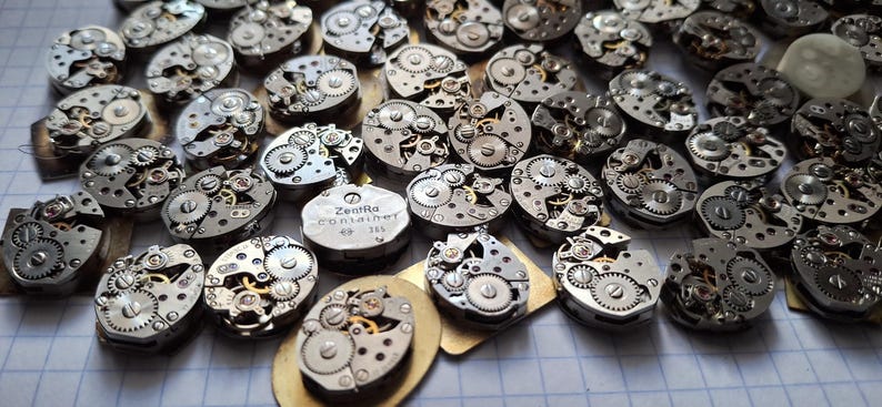 May include: A collection of vintage watch movements, showcasing intricate gears and mechanisms. The silver-toned components are arranged on a light-colored surface, with a few gold-toned bases. One movement has the text "Zentra Container 365".
