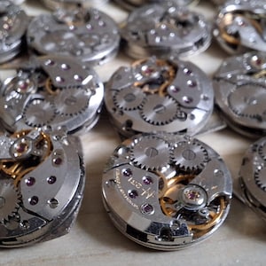 May include: A collection of disassembled watch movements, showcasing intricate gears, springs, and other mechanical components. The silver-toned metal parts are arranged in a grid pattern, with hints of gold and purple accents.