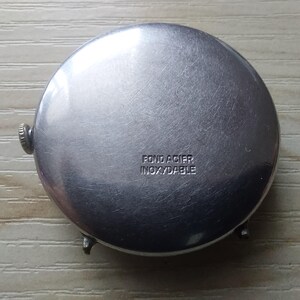 Puede incluir: Primer plano de la parte trasera de un reloj plateado. La parte trasera del reloj es redonda con un acabado de metal cepillado. El texto "FOND ACIER INOXYDABLE" est&aacute; estampado en la parte trasera del reloj. El reloj tiene una peque&ntilde;a corona y dos asas.