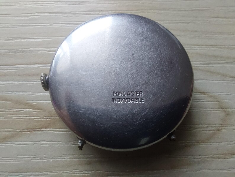 Puede incluir: Primer plano de la parte trasera de un reloj plateado. La parte trasera del reloj es redonda con un acabado de metal cepillado. El texto "FOND ACIER INOXYDABLE" est&aacute; estampado en la parte trasera del reloj. El reloj tiene una peque&ntilde;a corona y dos asas.