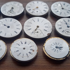 May include: A collection of antique watch faces, each with a white dial and black Roman numeral hour markers. Some faces have sub-dials. The watch faces are in various states of wear, with some showing signs of age and damage. The collection is arranged on a wooden surface.