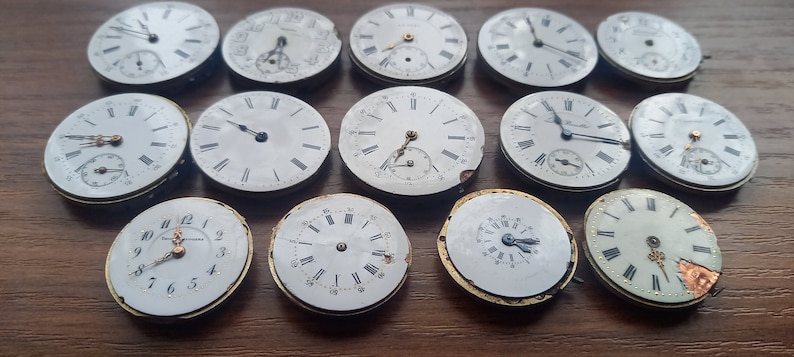 May include: A collection of antique watch faces, each with a white dial and black Roman numeral hour markers. Some faces have sub-dials. The watch faces are in various states of wear, with some showing signs of age and damage. The collection is arranged on a wooden surface.
