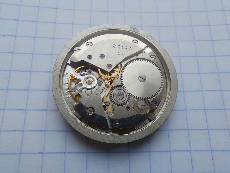 May include: A close-up view of a watch movement with intricate gears, springs, and a balance wheel. The metal components are silver and gold-toned, with the text "2414a SU" visible. The watch mechanism is set against a grid background.