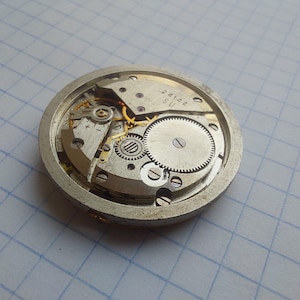 May include: Close-up of a vintage watch movement, showcasing intricate gears, springs, and a balance wheel. The silver-toned metal components are arranged within a circular frame. The text "2414A SU" is visible on a small plate.