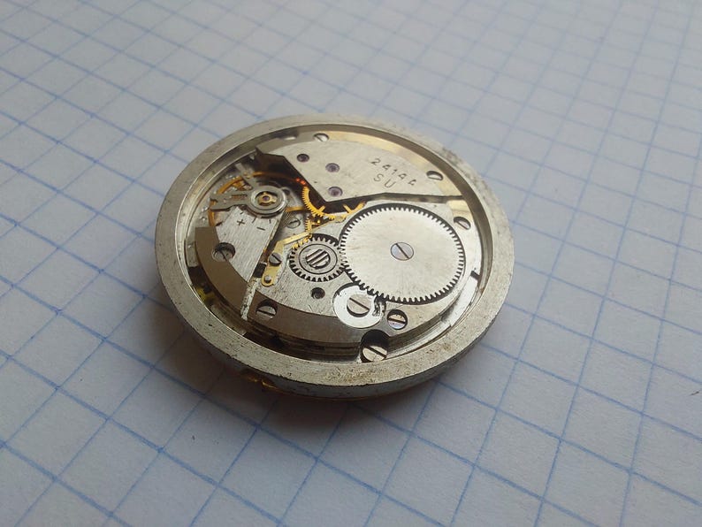 May include: Close-up of a vintage watch movement, showcasing intricate gears, springs, and a balance wheel. The silver-toned metal components are arranged within a circular frame. The text "2414A SU" is visible on a small plate.