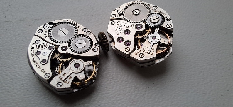 Peut inclure: Deux mouvements de montre Bulova vintage, pr&eacute;sentant des engrenages, des bijoux et des composants m&eacute;caniques complexes. Les m&eacute;canismes argent&eacute;s sont marqu&eacute;s "Bulova Watch Co. USA" et "21 Jewels". L'image met en &eacute;vidence la pr&eacute;cision de l'horlogerie.
