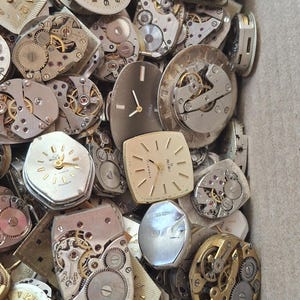 K&ouml;nnte beinhalten: Eine Sammlung von Vintage-Uhrenteilen, darunter Zifferbl&auml;tter, Zahnr&auml;der und Mechanismen. Die Zifferbl&auml;tter haben verschiedene Formen, darunter rund, quadratisch und rechteckig. Die Zahnr&auml;der sind aus Metall und haben komplizierte Designs. Die Teile befinden sich in einem Karton.